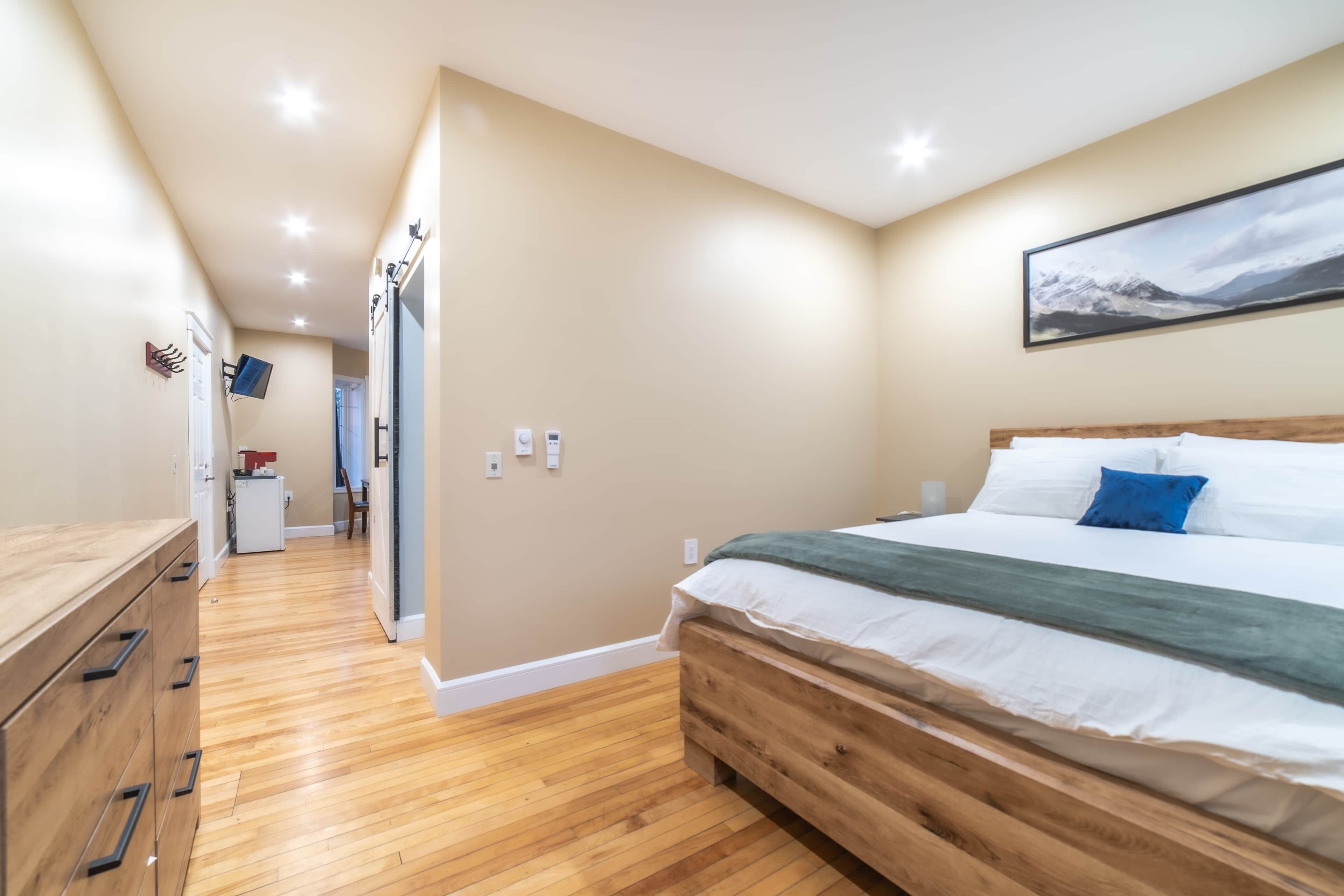 A bedroom with a wooden bed, a desk, and a chair. There is a hallway leading to a bathroom.