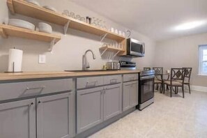 A kitchen with grey cabinets, a wooden countertop, a microwave, and a dining area with chairs.