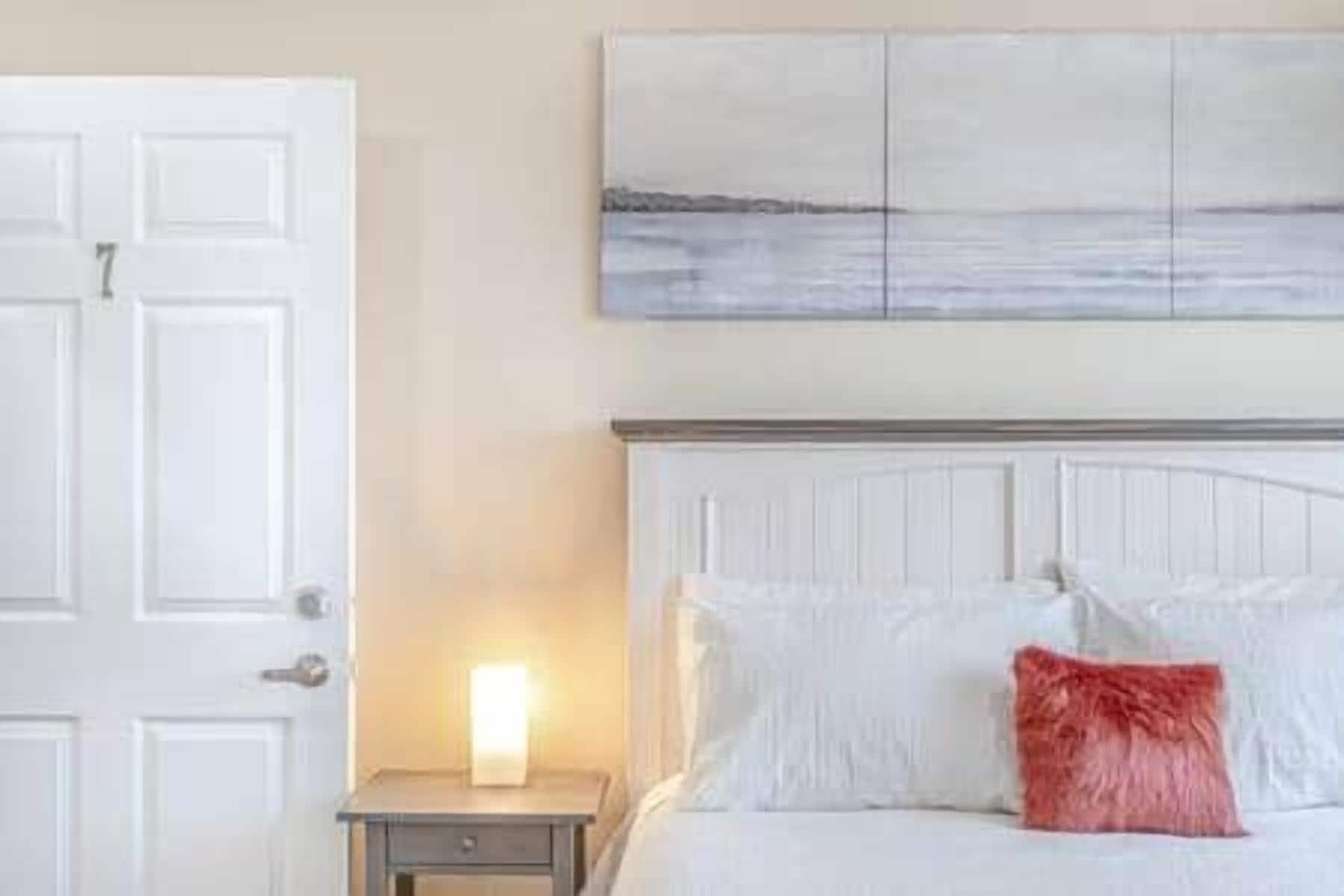 Quarto com cama branca, mesa de cabeceira, abajur e um quadro na parede.