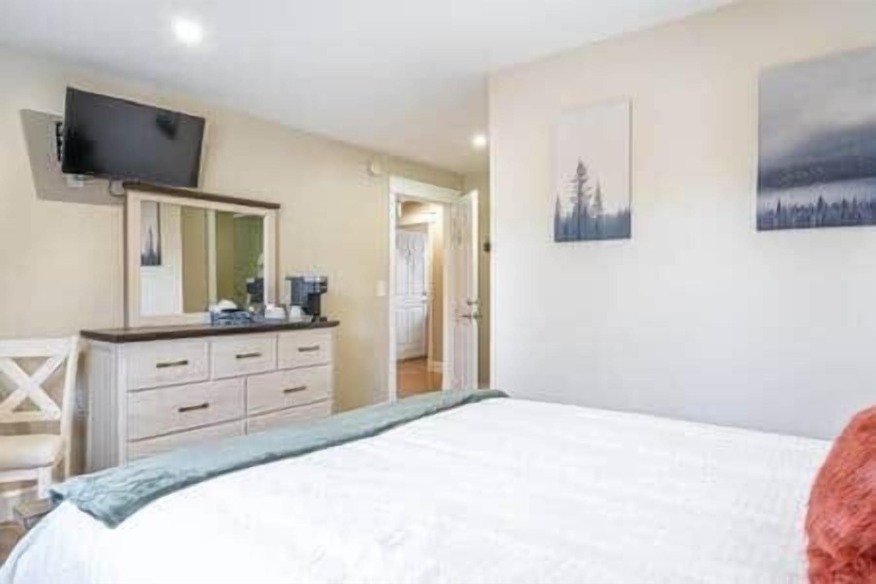 Un dormitorio con cama, cómoda, televisor montado en la pared y puerta que da a otra habitación.