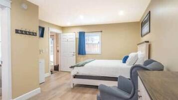 A bedroom with a bed, a desk, and a chair. There is a window with curtains, a door, and a wall-mounted sign.