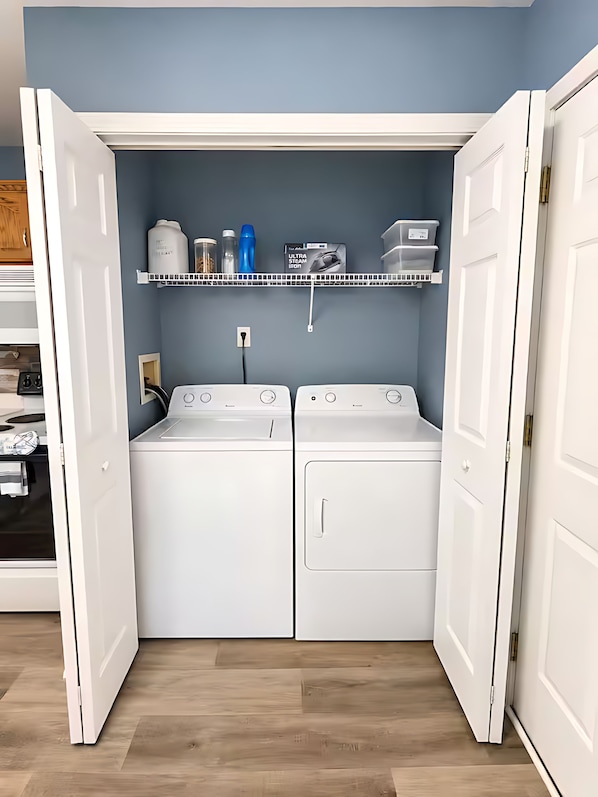 A laundry room with two white washing machines, a shelf above with storage containers, and a power outlet.