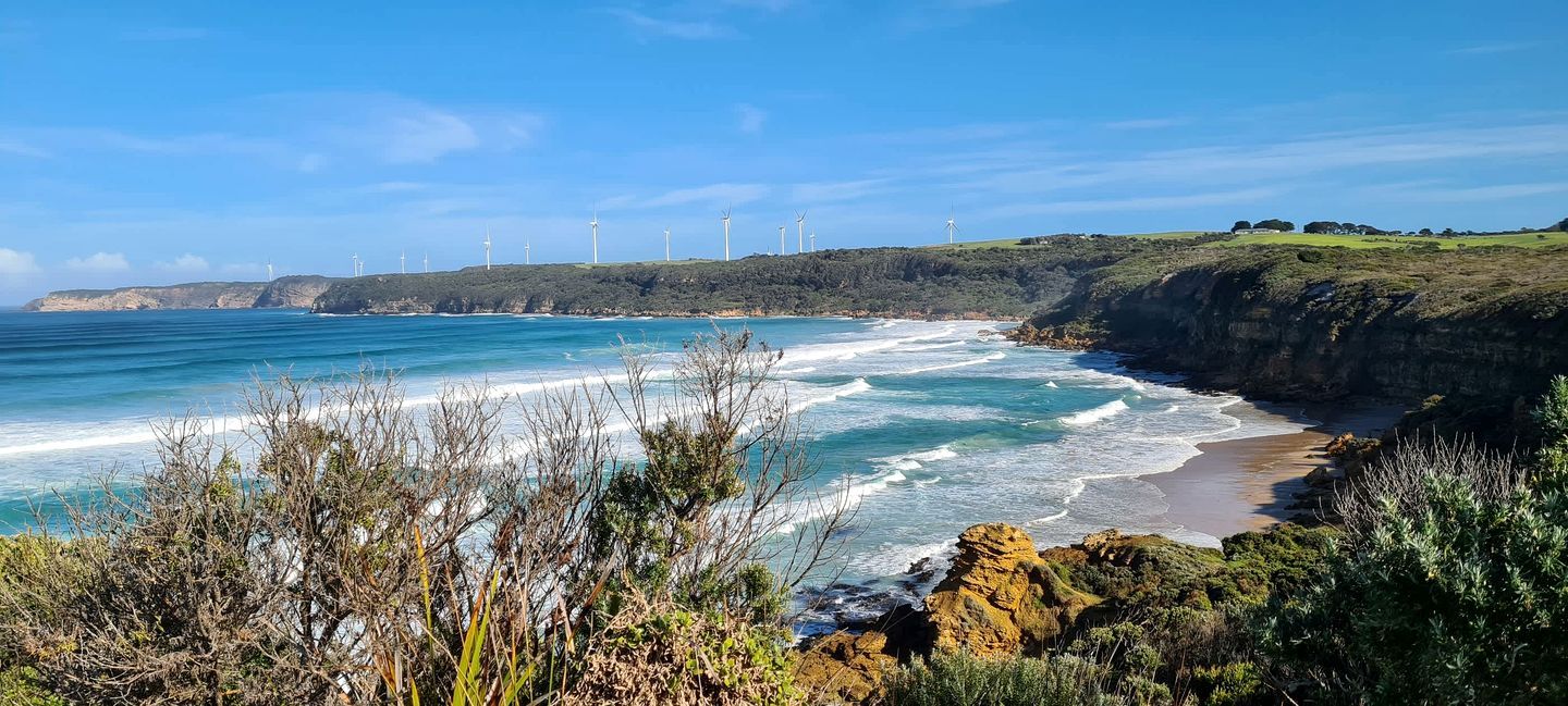 Eine Küstenlandschaft mit Strand, Meereswellen und Windkraftanlagen in der Ferne.