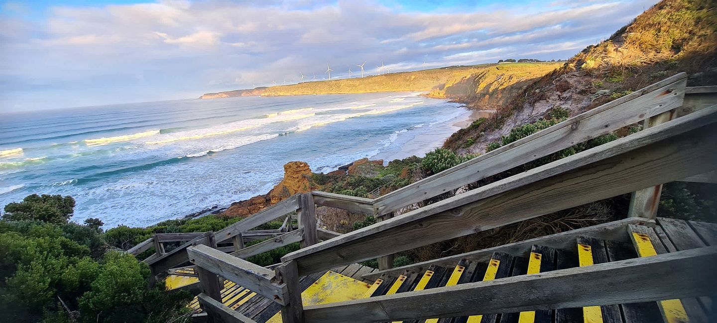 A coastal view with a wooden staircase leading down to the beach, overlooking the ocean and wind turbines in the distance.