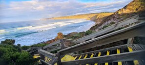 A coastal view with a wooden staircase leading down to the beach, overlooking the ocean and wind turbines in the distance.