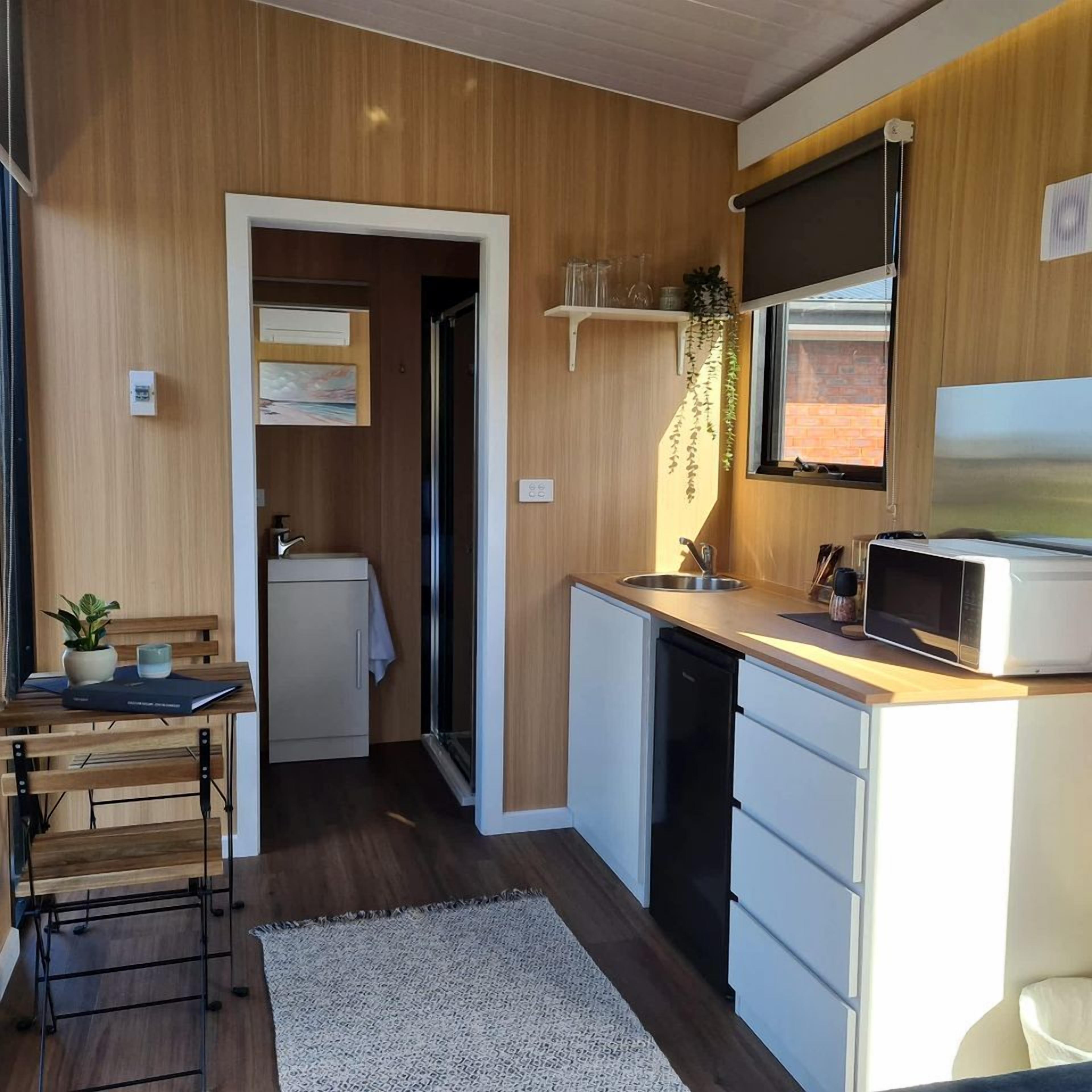 A compact kitchen with a microwave, sink, and a small refrigerator. There is a wooden bench with a plant and a book, and a white rug on the floor.