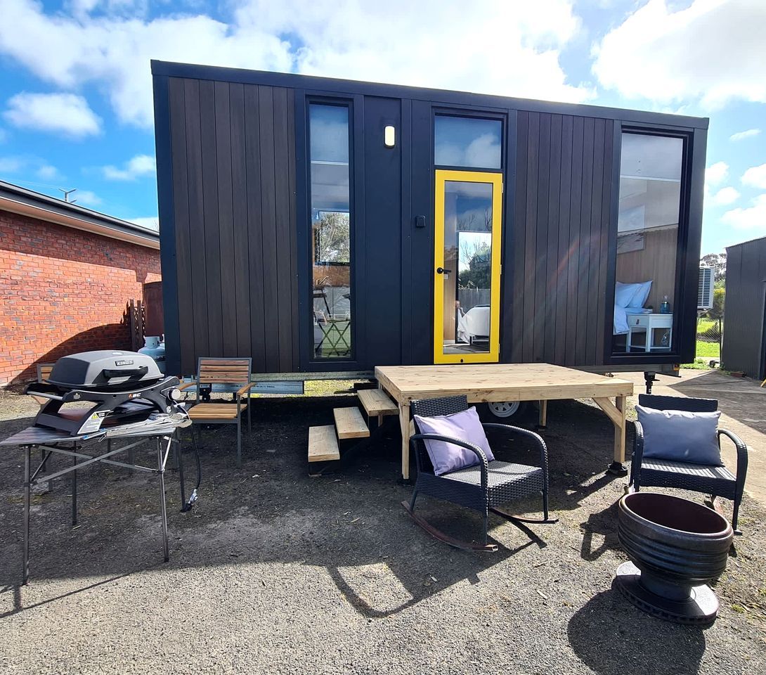 A modern outdoor patio with a wooden table and chairs, a barbecue grill, and a large window with a yellow door.