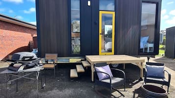 A modern outdoor patio with a wooden table and chairs, a barbecue grill, and a large window with a yellow door.