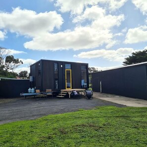 A modern, compact building with a flat roof, a door with a yellow frame, and a small outdoor seating area with a table and chairs.