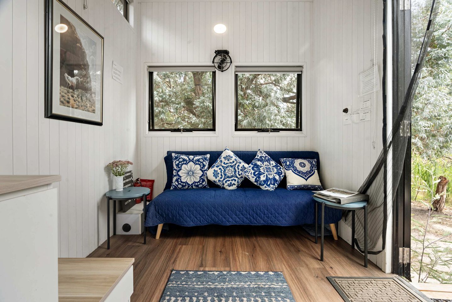 A room with a blue sofa, patterned cushions, a small table with a vase, and two windows with a view of trees.