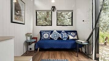 A room with a blue sofa, patterned cushions, a small table with a vase, and two windows with a view of trees.