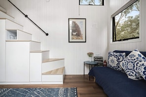A room with a staircase, a bed with patterned pillows, a framed picture on the wall, and a small table with a vase.