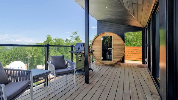 A wooden deck with a grill, a round wooden sauna, and a seating area with a table and chairs.