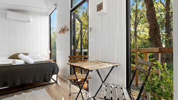 A bedroom with a bed, a small table, and a view of the outdoors through large windows.