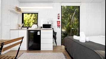Une pièce compacte comprenant un lit, une petite kitchenette, une fenêtre donnant sur un espace verdoyant et un extincteur fixé au mur.