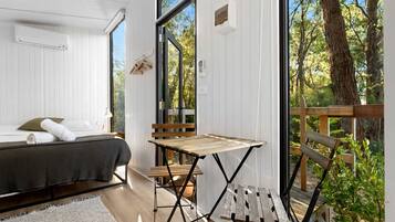 A bedroom with a bed, a small table, and a view of the outdoors through large windows.