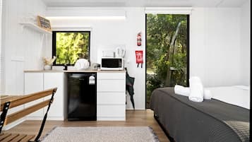 A compact room with a bed, a small kitchenette, a window with a view of greenery, and a fire extinguisher on the wall.