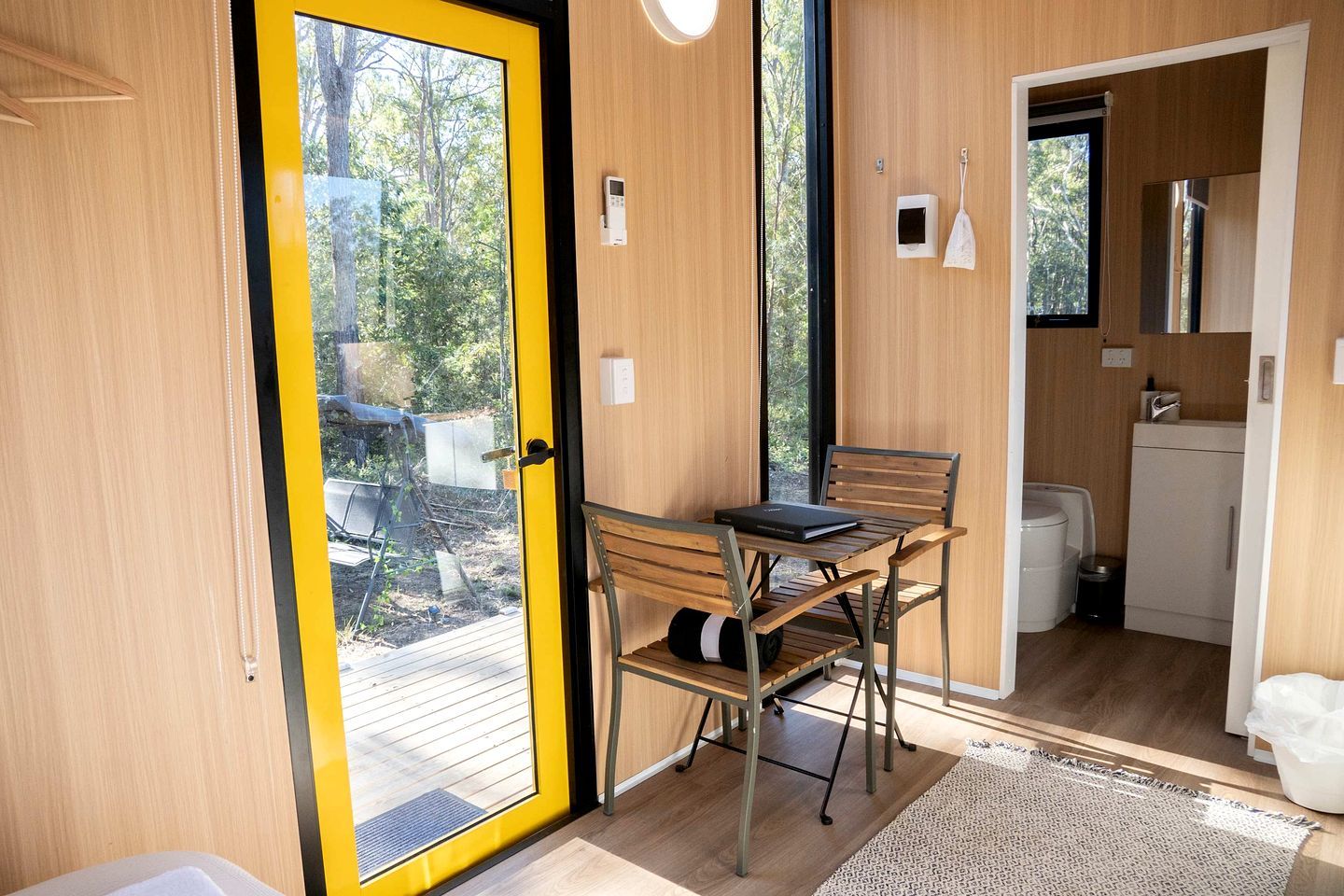 A compact room with a wooden floor, a small table with chairs, a window with a yellow frame, and a view of the outdoors.