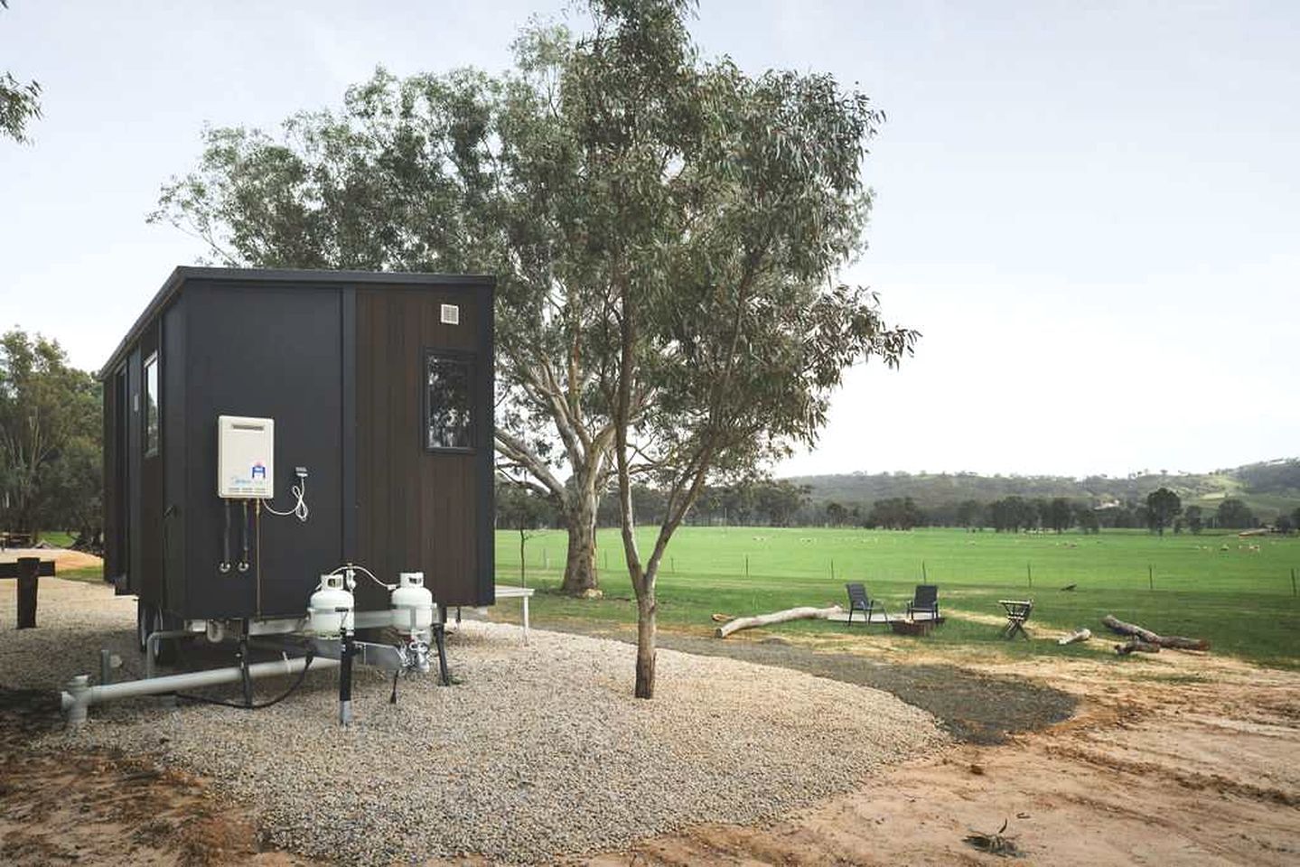 A small, dark-colored utility building with a water tank and pipes, located in a rural setting with a gravel ground and a grassy field in the background.