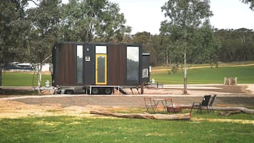 Una casa móvil pequeña y de color oscuro con una puerta amarilla, ubicada en un área cubierta de césped con árboles al fondo.