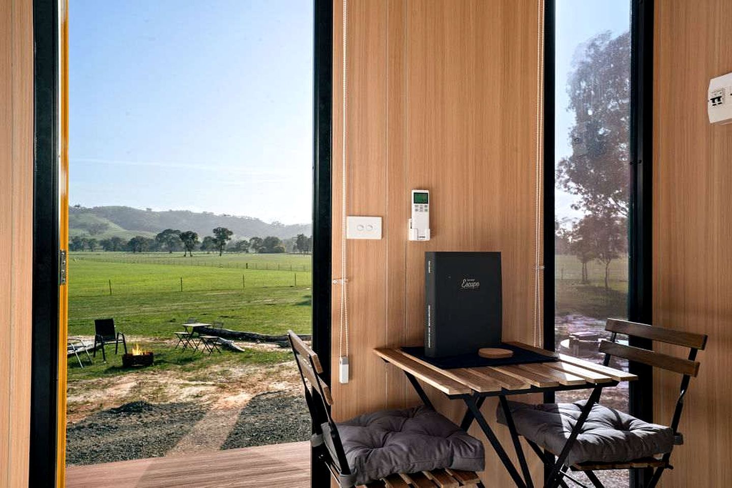 A room with a view of a green field, a table with chairs, a fire pit, and a black box on the wall.
