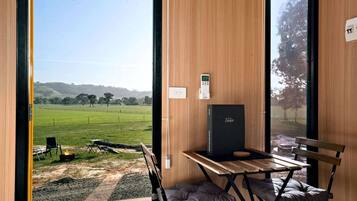 A room with a view of a green field, a table with chairs, a fire pit, and a black box on the wall.