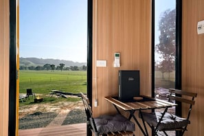 A room with a view of a green field, a table with chairs, a fire pit, and a black box on the wall.
