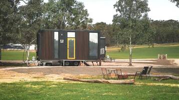 Una casa móvil pequeña y de color oscuro con una puerta amarilla, ubicada en un área cubierta de césped con árboles al fondo.