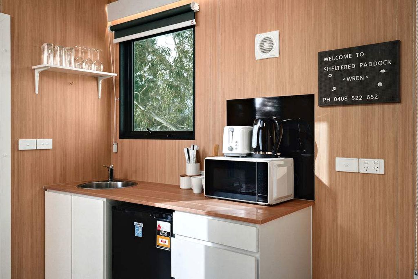Een compacte keuken met een magnetron, broodrooster en koffiezetapparaat, een raam met uitzicht op bomen, en een welkomstbord.