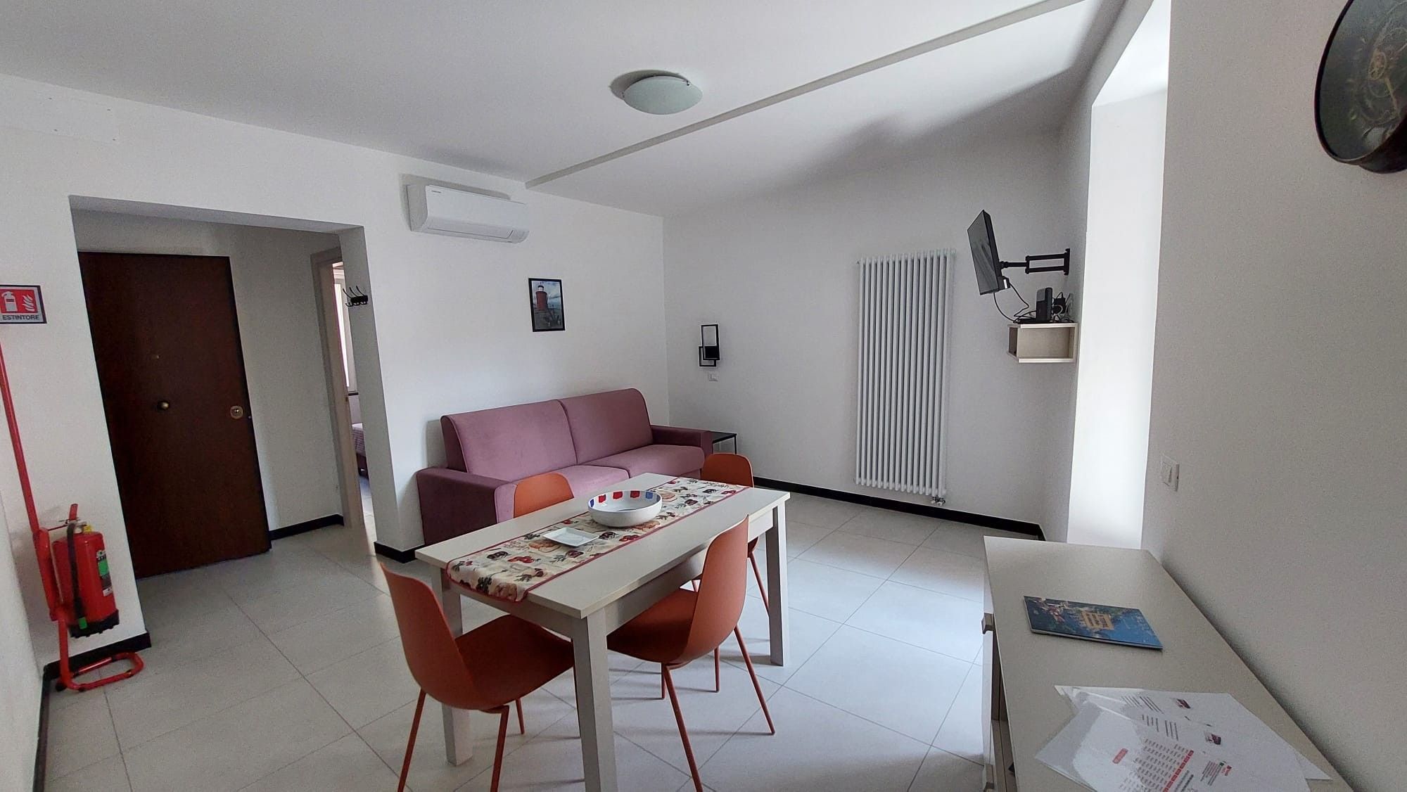 Ett kompakt bostadsutrymme med ett matbord, två stolar, en soffa, ett element och en brandsläckare.