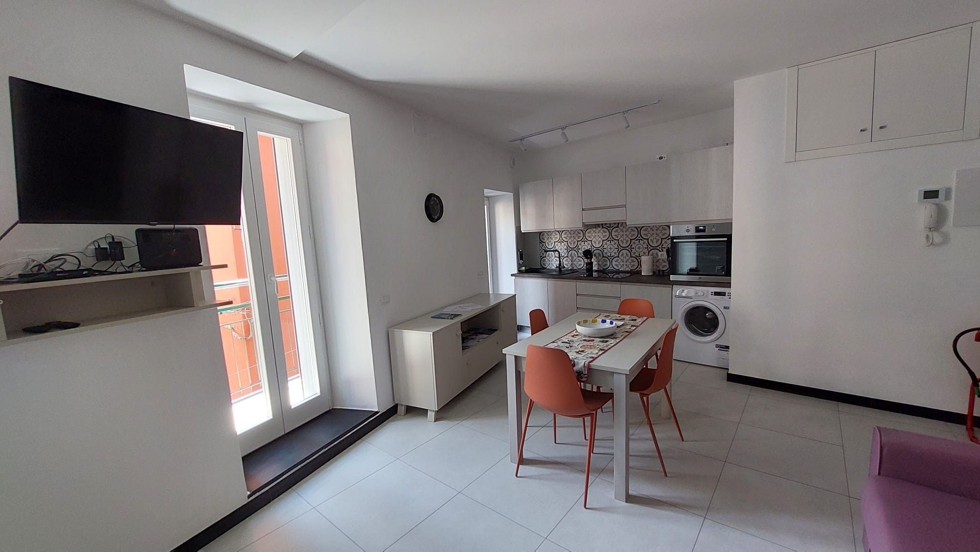 A modern kitchen with a dining area, a washing machine, and a flat-screen TV mounted on the wall.