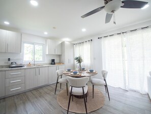 Una cocina moderna con armarios blancos, una mesa de comedor con sillas, un ventilador de techo y una ventana con cortinas.