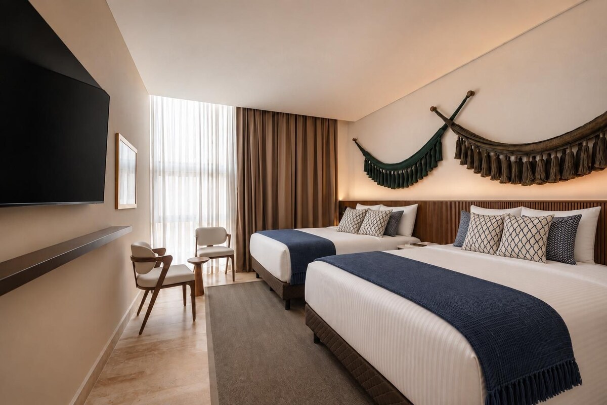A hotel room with two beds, a flat-screen TV mounted on the wall, a seating area with a chair and a small table, and a large window with curtains.