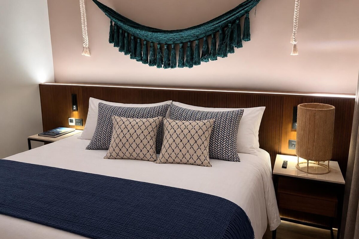 A neatly made bed with patterned pillows, a blue throw blanket, and a hammock hanging from the ceiling.