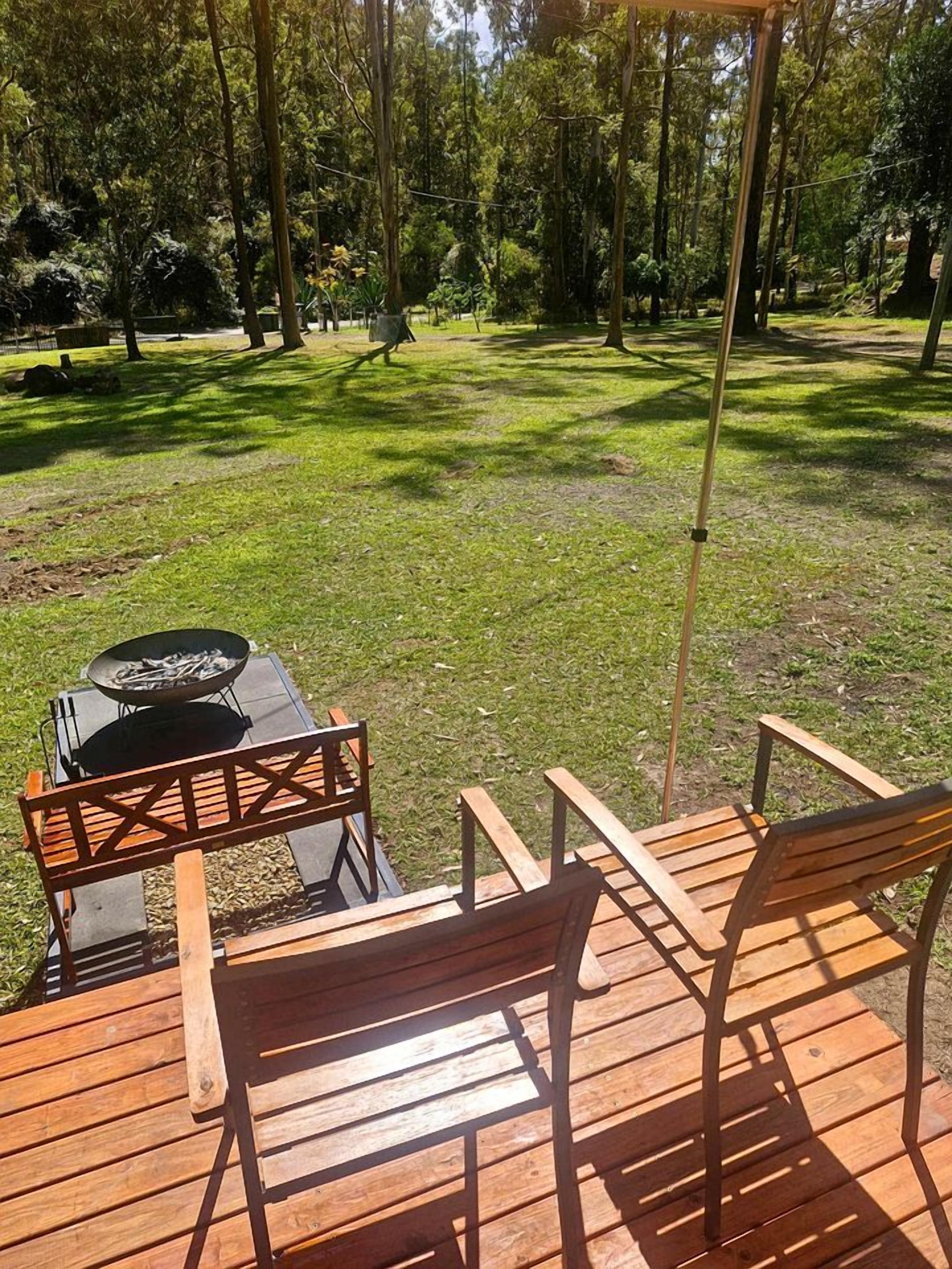 Um deck de madeira ao ar livre, com uma mesa e cadeiras, rodeado por uma área gramada com árvores.