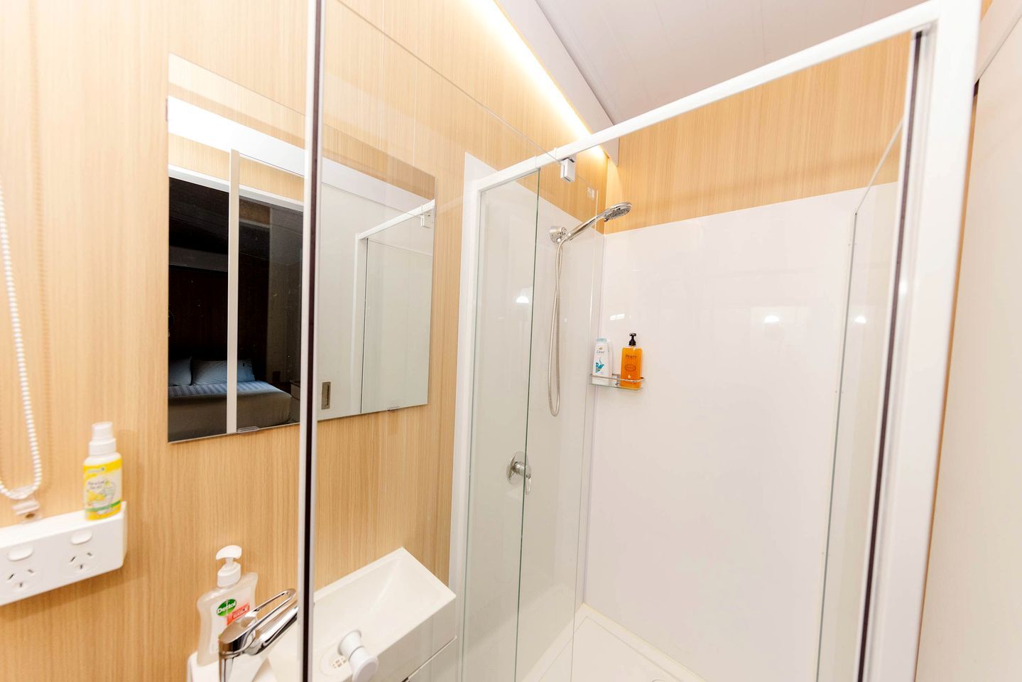 Ein modernes Badezimmer mit einer gläsernen Duschkabine, einem Spiegel, einem Waschbecken mit Seifenspender und einer Shampoo-Flasche.