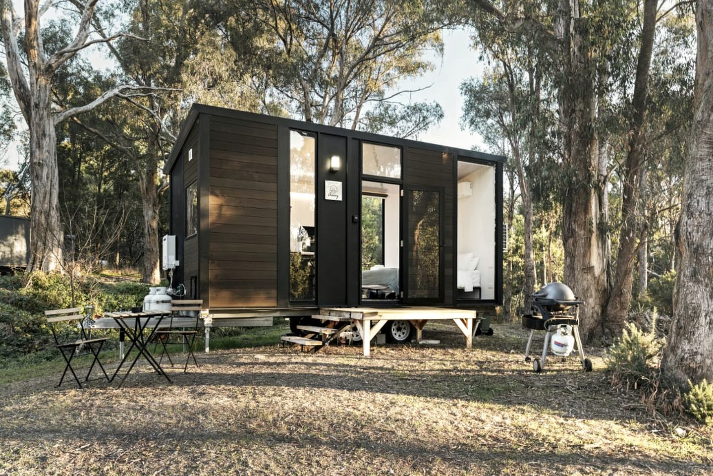 Eine kleine Holzhütte mit einem Esstisch und Stühlen im Freien, einem Grill und einer Transporttoilette.
