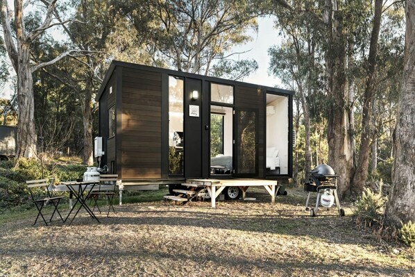 Una cabaña pequeña con una mesa y sillas en el exterior, una parrilla y un baño químico.