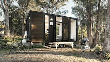 A small cabin with a dining table and chairs outside, a grill, and a portable toilet.