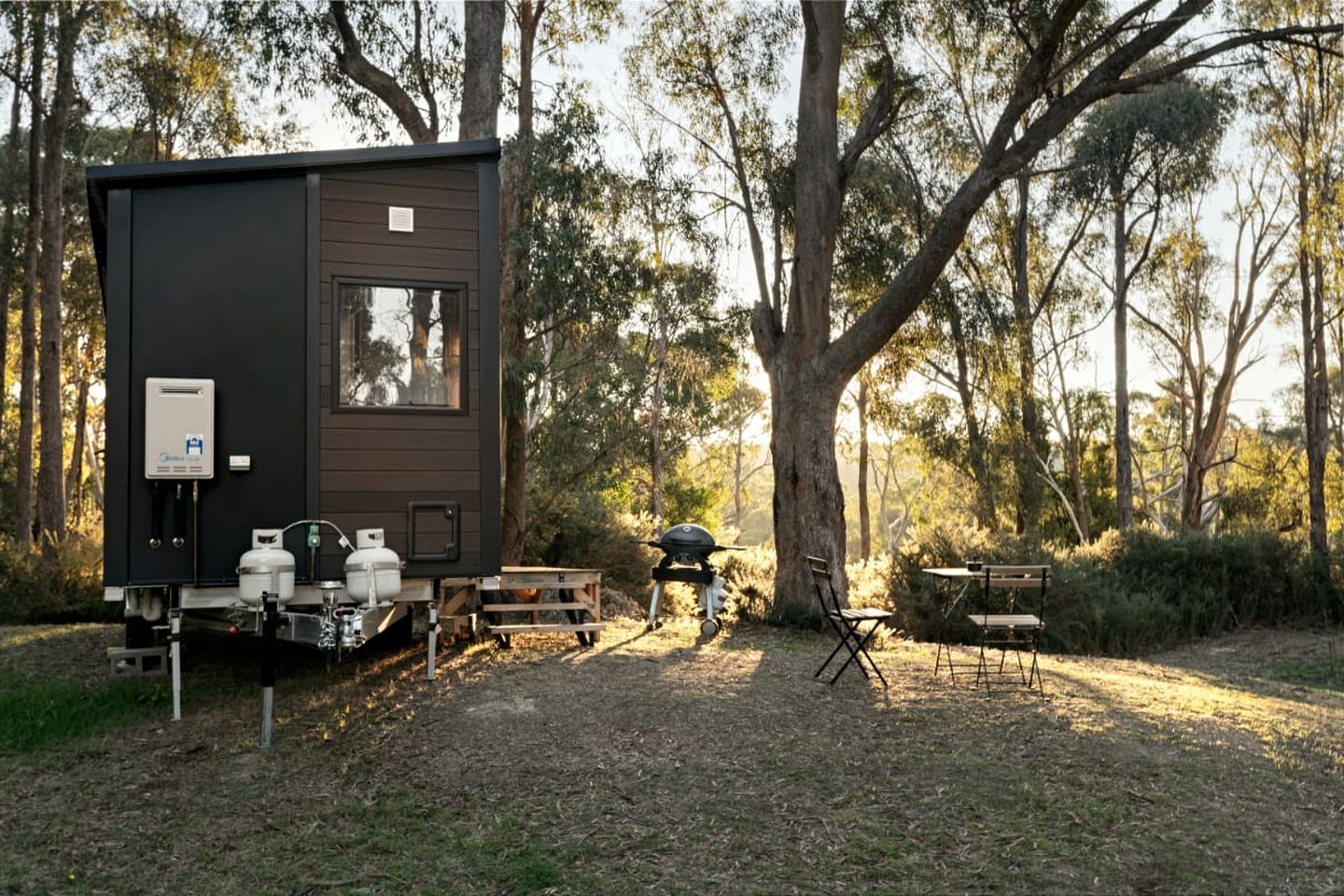 A compact, dark-colored RV with a water heater and propane tanks, set in a wooded area with a barbecue grill and foldable chairs.