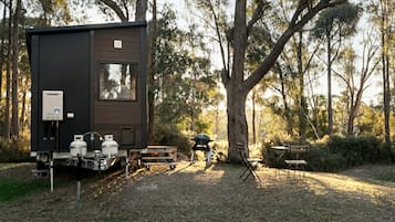 A compact, dark-colored RV with a water heater and propane tanks, set in a wooded area with a barbecue grill and foldable chairs.