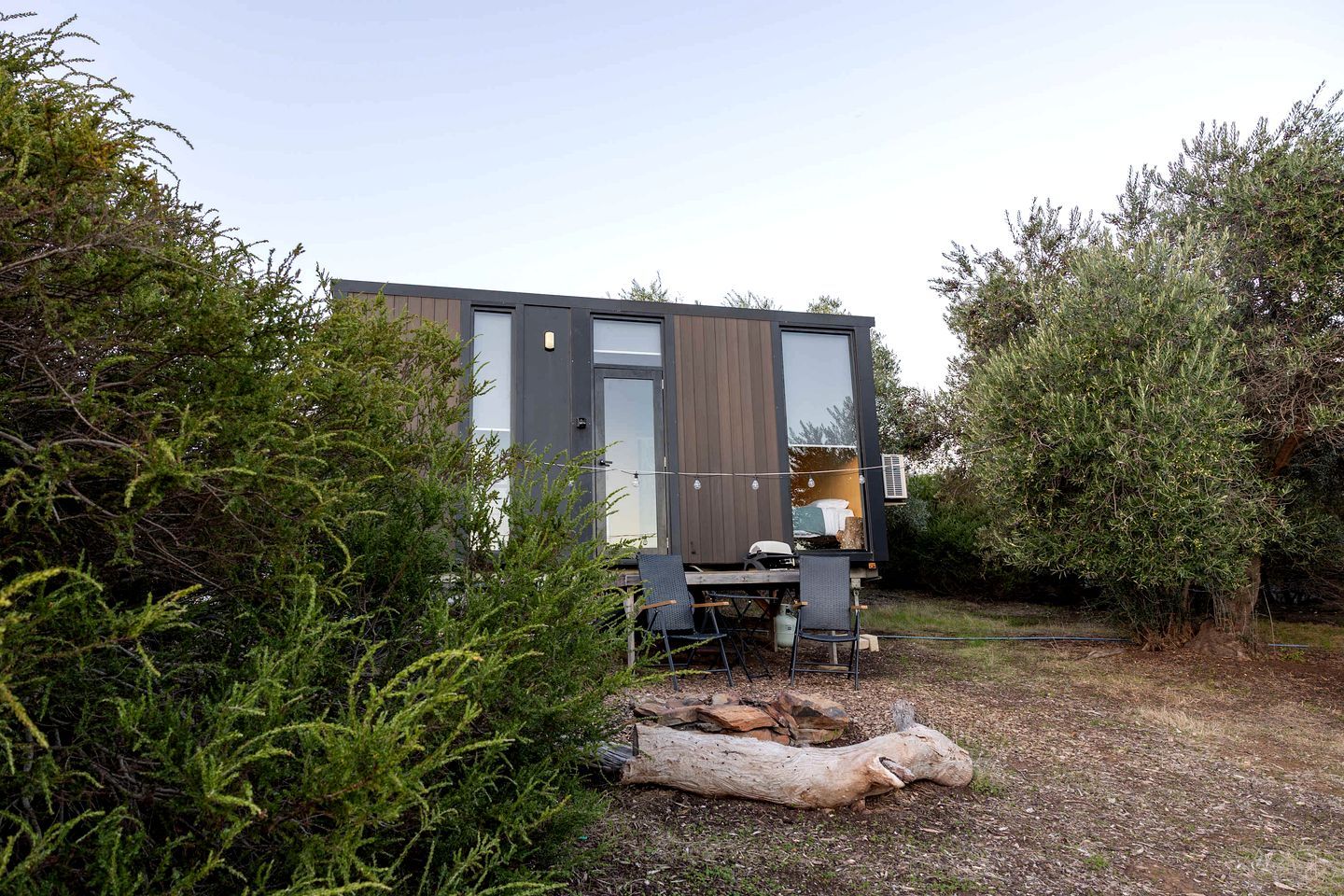 A modern, compact mobile home with a large window, a dining area with chairs, and a fire pit.