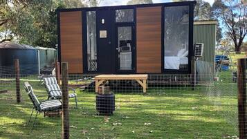 A small cabin with a door, a window, and a bed inside. There is a table and chairs outside.