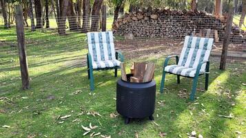 Twee stoelen met daar tussenin een vuurkorf, op een gazon met een hek en bomen op de achtergrond.