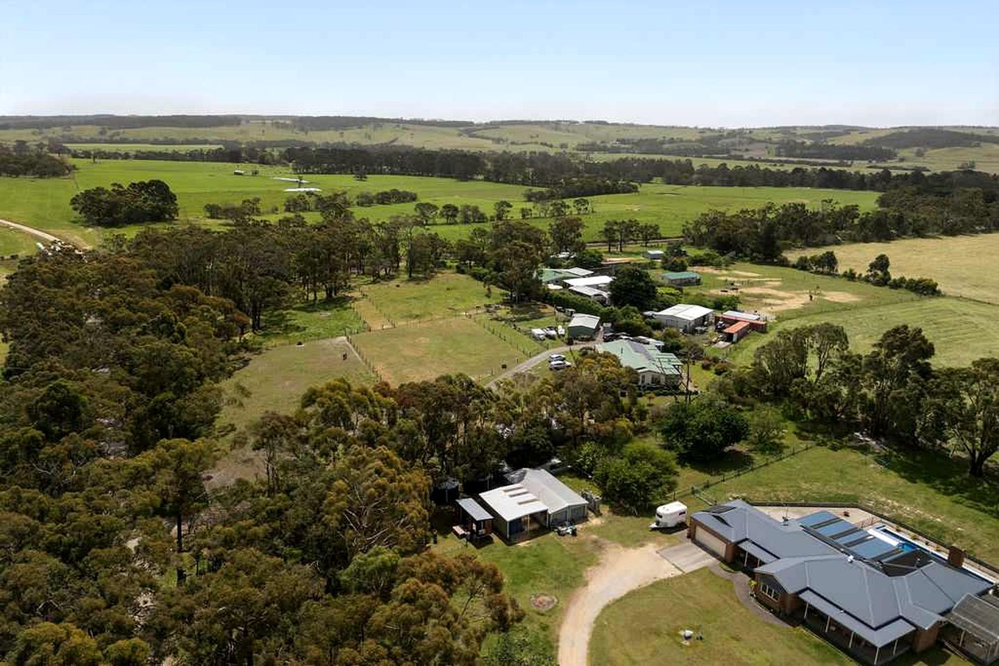 An aerial view of a rural property with multiple buildings, including a large house with a sloped roof and a smaller structure with a flat roof, surrounded by open fields and scattered trees.