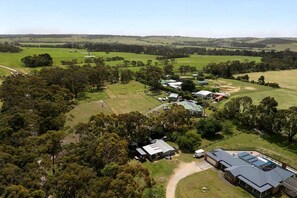 An aerial view of a rural property with multiple buildings, including a large house with a sloped roof and a smaller structure with a flat roof, surrounded by open fields and scattered trees.