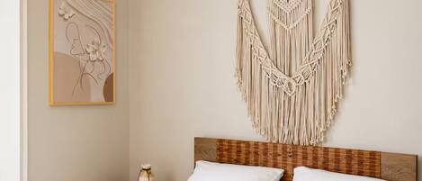 Una cama bien hecha con sábanas blancas, cabecera de madera y un colgador de macramé en la pared.