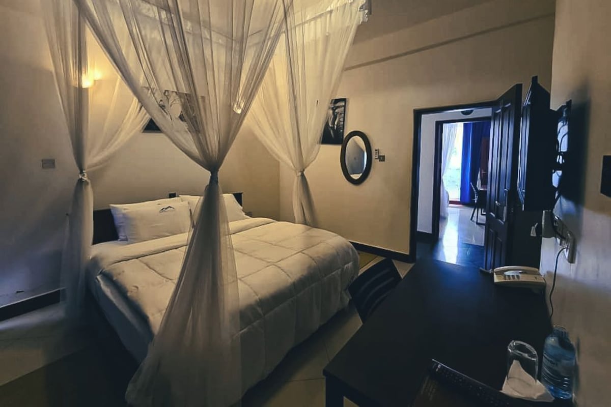 A hotel room with a bed, a desk, a chair, a bedside table, a mirror, and a door leading to another room.