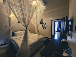 Habitación de hotel con cama, escritorio, silla, mesita de noche, espejo y puerta que da a otra habitación.