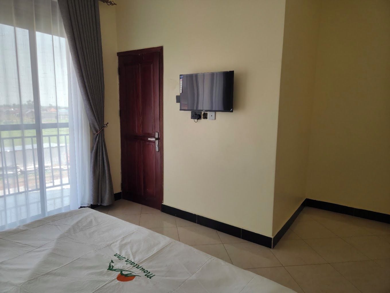 Ein Zimmer mit einem Bett, einer Tür, einem an der Wand befestigten Fernseher und einem Fenster mit Vorhängen.